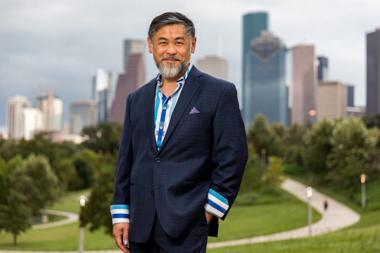 image of a male-identified figure with graying black hair and a salt and pepper beard dressed in a blue suit with a white and blue striped shirt. In the background is the Houston skyline.