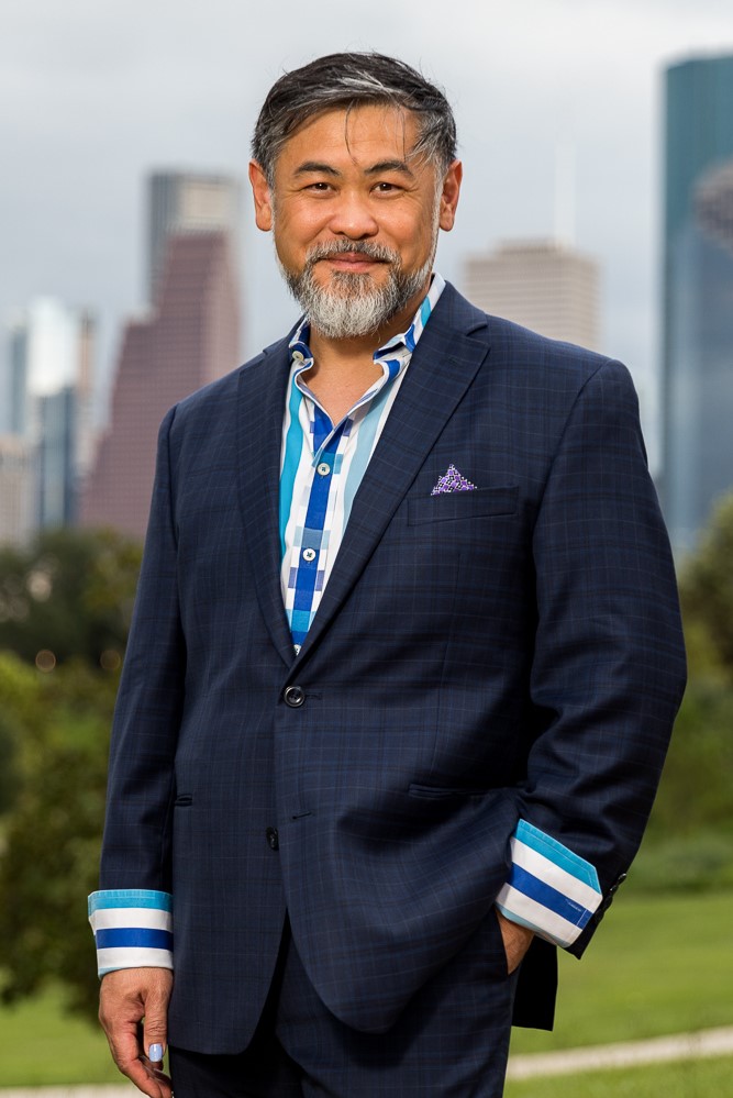 image of a male-identified figure with graying black hair and a salt and pepper beard dressed in a blue suit with a white and blue striped shirt. In the background is the Houston skyline.